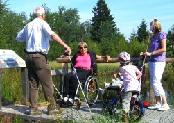 Holzsteg mit Infotafel im Hohen Venn. Auf dem Holzsteg stehen eine Rollstuhlfahrerin, ein Mann mit Wanderstock, eine Frau mit Kinderwagen und ein kleines Mädchen mit Fahrrad.
