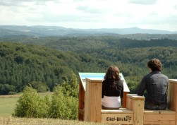 Ein Mann und eine Frau, die sich auf einer Holzbank mit der Aufschrift "Eifel-Blicke" ausruhen und den Blick genießen. Vor ihnen befindet sich eine Infotafel.